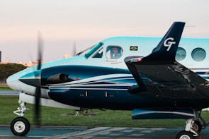 A turquoise and white twin-engine propeller aircraft with a person visible in the cockpit, parked on a tarmac. The Brazilian flag and an airline logo are displayed near the cockpit window, with blurred propellers indicating slight motion. In the background, there is a hint of an urban landscape and a green grass field.