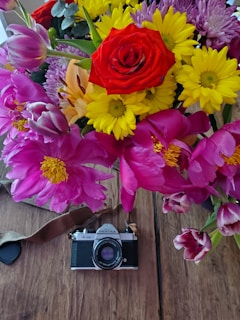 A vibrant bouquet of various flowers including a prominent red rose, yellow daisies, pink peonies, and purple tulips is arranged above a vintage Pentax camera. The flowers and the camera are set on a wooden surface, creating a contrasting blend of nature and nostalgia.