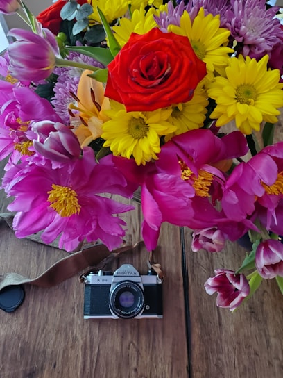 A vibrant bouquet of various flowers including a prominent red rose, yellow daisies, pink peonies, and purple tulips is arranged above a vintage Pentax camera. The flowers and the camera are set on a wooden surface, creating a contrasting blend of nature and nostalgia.