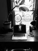 A workspace with a wooden desk featuring a laptop, glasses, a pen, and a drink. A potted plant and dreamcatchers hang in a window behind, casting silhouettes and creating an artistic, cozy atmosphere.