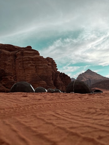 A desert landscape featuring a series of geodesic dome tents set against the backdrop of large, rugged rock formations. The sky above is partially cloudy, adding to the vast and open feeling of the scene.