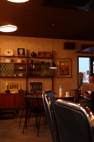 A cozy vintage-themed cafe featuring wooden shelves with various decorative items, including artwork and pottery. Retro signs labeled ‘KAFFE’ and ‘KARSK’ are displayed. A person is seated at a table, using a smartphone, with dim lighting contributing to the warm ambiance.