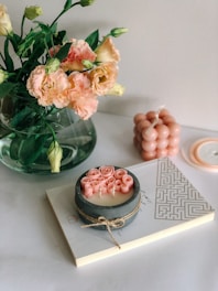 A bouquet of pink and cream carnations with green leaves is placed in a clear vase. Next to it, there is a decorative candle with a rustic twine wrap featuring rose-shaped embellishments. The candle is resting on a book with a geometric pattern on the cover. A sculpted bubble candle in pastel pink is also visible, along with part of a round lid.