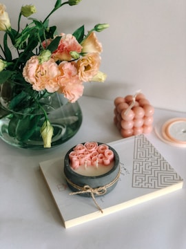 A bouquet of pink and cream carnations with green leaves is placed in a clear vase. Next to it, there is a decorative candle with a rustic twine wrap featuring rose-shaped embellishments. The candle is resting on a book with a geometric pattern on the cover. A sculpted bubble candle in pastel pink is also visible, along with part of a round lid.