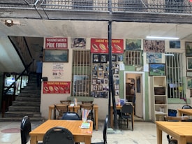 An interior space resembling a traditional eatery with tables and chairs neatly arranged. Walls are adorned with numerous framed photographs and paintings displaying various scenes, suggesting a cultural or historical theme. Colorful straws are placed in containers on the tables. Signage in a foreign language is visible, along with menus and an entryway leading to another room. The atmosphere appears casual and inviting.