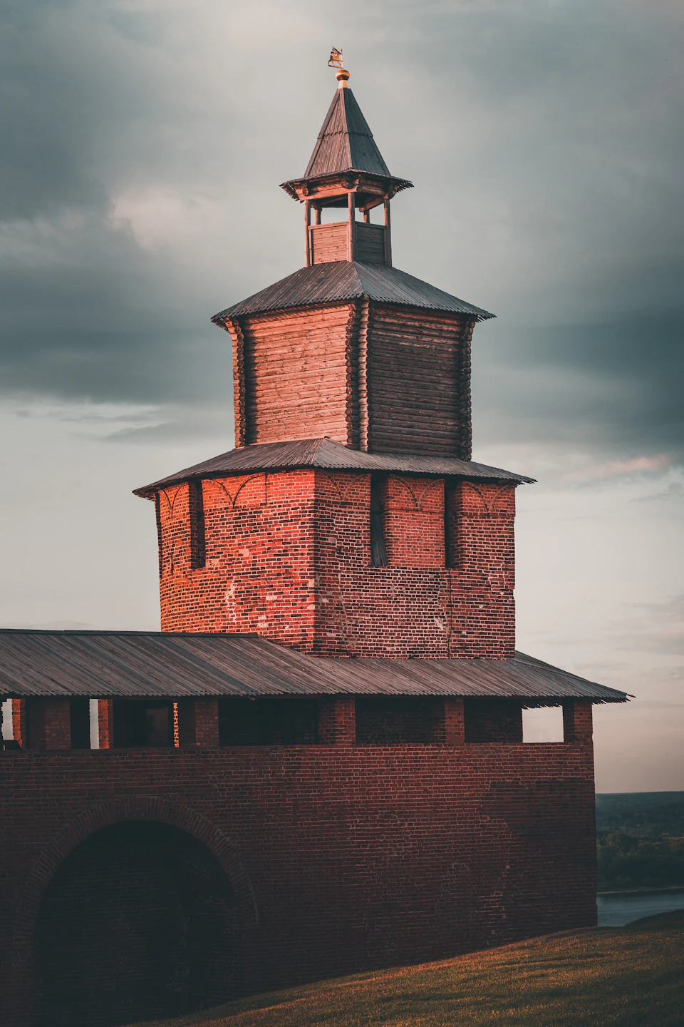The image depicts a tall brick tower with a pointed roof and a small dome at the top, standing against an overcast sky. The tower is situated on a hillside, surrounded by lush green grass that contrasts with its reddish-brown hue. In the foreground, there's a large archway leading up to the tower, adding depth to the scene.