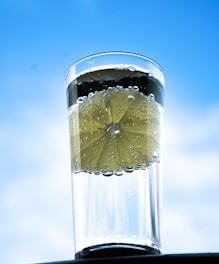 A tall glass filled with clear sparkling water and a slice of lemon immersed in it, with bubbles clinging to the lemon slice. The background is a bright, clear blue sky, giving the image a fresh and invigorating feel.
