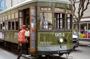 A green vintage streetcar labeled 954 on the front travels on a street. A person wearing a red baseball cap and a red and white jacket is boarding the streetcar. The streetcar displays a sign for St. Charles and has one headlight. Trees and building storefronts are visible in the background.