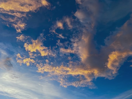 A sky filled with fluffy clouds, illuminated by a warm golden hue, set against a backdrop of deep blue. The clouds are scattered in various formations, with some appearing dense and others more wispy, creating a dynamic and visually pleasing scene.