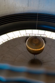 A large brass pendulum suspended in a circular room, swinging over a stone floor marked with numbers. The numbered floor forms a spiral-like pattern. The room has an industrial feel with concrete walls and a circular light fixture surrounding the pendulum at the upper part.