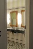 A bathroom interior featuring two sinks with ornate mirrors framed in gold. Wall-mounted lights are positioned vertically beside each mirror. The walls are covered in white tiles, and the floor has a patterned mosaic design. Plumbing and paper towel holders are visible under the sinks.