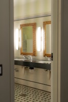 A bathroom interior featuring two sinks with ornate mirrors framed in gold. Wall-mounted lights are positioned vertically beside each mirror. The walls are covered in white tiles, and the floor has a patterned mosaic design. Plumbing and paper towel holders are visible under the sinks.