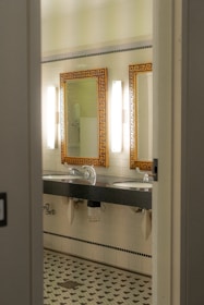 A bathroom interior featuring two sinks with ornate mirrors framed in gold. Wall-mounted lights are positioned vertically beside each mirror. The walls are covered in white tiles, and the floor has a patterned mosaic design. Plumbing and paper towel holders are visible under the sinks.