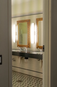 A bathroom interior featuring two sinks with ornate mirrors framed in gold. Wall-mounted lights are positioned vertically beside each mirror. The walls are covered in white tiles, and the floor has a patterned mosaic design. Plumbing and paper towel holders are visible under the sinks.