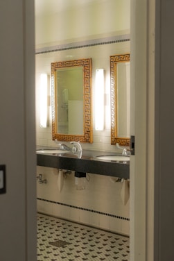 A bathroom interior featuring two sinks with ornate mirrors framed in gold. Wall-mounted lights are positioned vertically beside each mirror. The walls are covered in white tiles, and the floor has a patterned mosaic design. Plumbing and paper towel holders are visible under the sinks.