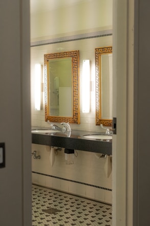 A bathroom interior featuring two sinks with ornate mirrors framed in gold. Wall-mounted lights are positioned vertically beside each mirror. The walls are covered in white tiles, and the floor has a patterned mosaic design. Plumbing and paper towel holders are visible under the sinks.