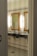 A bathroom interior featuring two sinks with ornate mirrors framed in gold. Wall-mounted lights are positioned vertically beside each mirror. The walls are covered in white tiles, and the floor has a patterned mosaic design. Plumbing and paper towel holders are visible under the sinks.