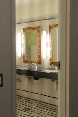 A bathroom interior featuring two sinks with ornate mirrors framed in gold. Wall-mounted lights are positioned vertically beside each mirror. The walls are covered in white tiles, and the floor has a patterned mosaic design. Plumbing and paper towel holders are visible under the sinks.