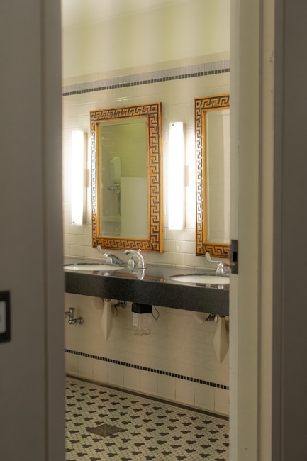 A bathroom interior featuring two sinks with ornate mirrors framed in gold. Wall-mounted lights are positioned vertically beside each mirror. The walls are covered in white tiles, and the floor has a patterned mosaic design. Plumbing and paper towel holders are visible under the sinks.