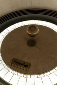 A large pendulum hanging from the ceiling in a circular room. The pendulum is suspended over a circular platform marked with numbers around its edge, possibly indicating time or degrees. The surroundings are minimalistic, with a few architectural elements visible on the periphery of the image.
