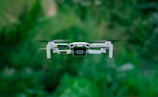 A small quadcopter drone with four propellers hovers in the foreground, featuring a camera mounted at its front. The drone's body is predominantly white and has the text 'MINI 2' visible on one of the arms. The background is filled with lush greenery, providing a natural setting.