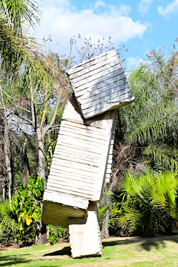 A tall, abstract sculpture made of stacked rectangular stone slabs stands in a lush, tropical garden. The slabs have a ridged texture and are precariously balanced, creating an impression of movement. Surrounding the sculpture, various green plants and palm trees contribute to the vibrant garden setting under a bright blue sky with patches of white clouds.