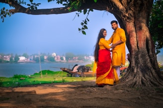 A couple stands together under a large, leafy tree by the side of a calm body of water. They are wearing traditional attire, with the woman in a vibrant red and yellow sari and the man in a matching yellow outfit. The background shows a serene riverside landscape with green grass, a boat, and houses across the water. Sunlight filters through the tree leaves, casting gentle shadows on the ground.
