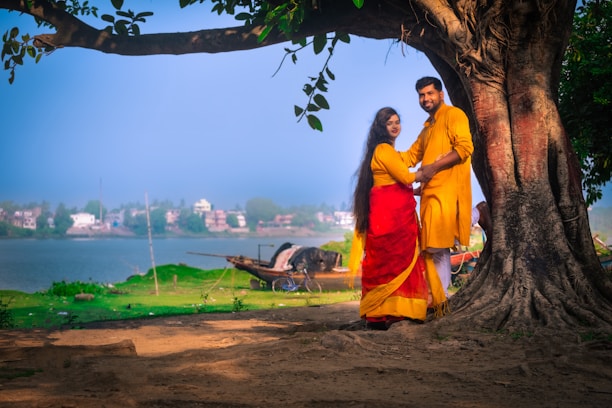 A couple stands together under a large, leafy tree by the side of a calm body of water. They are wearing traditional attire, with the woman in a vibrant red and yellow sari and the man in a matching yellow outfit. The background shows a serene riverside landscape with green grass, a boat, and houses across the water. Sunlight filters through the tree leaves, casting gentle shadows on the ground.