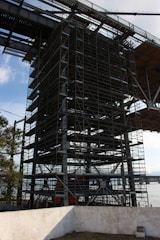 A large structure featuring extensive scaffolding and metal beams, situated near a body of water. The scaffolding is complex, indicating construction or maintenance activity. A concrete wall is visible in the foreground, and there are trees nearby. The sky is partly cloudy, indicating a clear day.