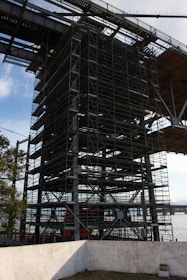 A large structure featuring extensive scaffolding and metal beams, situated near a body of water. The scaffolding is complex, indicating construction or maintenance activity. A concrete wall is visible in the foreground, and there are trees nearby. The sky is partly cloudy, indicating a clear day.