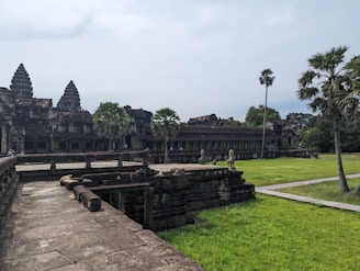 An ancient stone temple complex is set against a cloudy sky, featuring intricately carved towers resembling lotus buds. Surrounding the temple are well-kept green lawns and a few tall palm trees. The architecture exhibits weathered stones indicating age and historical significance.