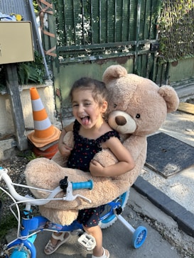 A young child, wearing a black dress with colorful polka dots, is happily sitting on a blue tricycle. The child is playfully sticking out their tongue while holding a large teddy bear. The setting is outdoors near a fenced gate. An orange and white traffic cone is visible in the background.