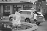 A black and white image featuring a large SUV parked near a building with Chinese signage. In the foreground, a child stands near the back of the vehicle holding something, possibly a snack. A fish-shaped sculpture is visible near a water feature, and another person stands in the background near the building entrance.