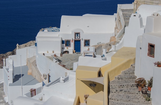 A serene coastal scene featuring traditional whitewashed buildings with blue accents, including doors and windows. The structures are built along a terraced hillside overlooking a vast, deep blue sea. Stone steps and pathways wind through the area, with scattered potted plants adding a touch of greenery.