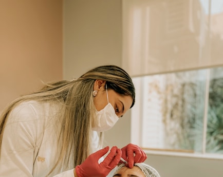 A woman wearing a white coat, face mask, and red gloves is administering an injection to a patient. The patient is lying down, partially visible, wearing a hairnet. Natural light filters through a large window, creating a calm atmosphere.
