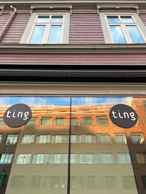 A storefront window reflects a large brick building with multiple windows under a clear sky. Above the window are decorative moldings and two signs on either side displaying the word 'ting' in white letters on a black circular background. The building's upper section features large windows with white frames against a brown, horizontal wooden panel siding.