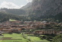 A picturesque village nestled in a lush mountainous landscape, with numerous traditional wooden houses spread across terraced land. The backdrop features towering green mountains, dense forests, and a slightly cloudy sky.