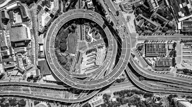 An aerial view captures a complex urban highway interchange with multiple layers of roads and overpasses forming intricate patterns. Vehicles move smoothly along the roads, which encircle a central area featuring green spaces and trees. Surrounding the interchange are densely packed buildings and structures, depicting a bustling city environment.