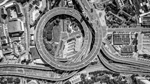 An aerial view captures a complex urban highway interchange with multiple layers of roads and overpasses forming intricate patterns. Vehicles move smoothly along the roads, which encircle a central area featuring green spaces and trees. Surrounding the interchange are densely packed buildings and structures, depicting a bustling city environment.