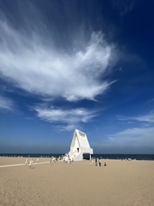 A modern, white chapel with a triangular roof stands on a sandy beach by the ocean. The sky above is bright blue with sweeping white clouds. People are walking around the chapel, enjoying the beach setting.