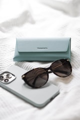 A light blue Tiffany & Co. glasses case is placed on a white textured surface, accompanied by a pair of dark sunglasses and a smartphone with a light-colored case.