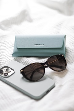 A light blue Tiffany & Co. glasses case is placed on a white textured surface, accompanied by a pair of dark sunglasses and a smartphone with a light-colored case.