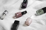 Several nail polish bottles in different colors are scattered on a textured white fabric background. The arrangement appears random, showcasing a variety of brands and shades.