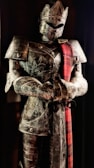 A full suit of medieval armor is displayed, featuring intricate engravings and a red plaid sash draped over the shoulder. The armor includes a helmet with a visor, breastplate, and gauntlets. It stands upright in a slightly darkened setting, giving an imposing and majestic appearance.