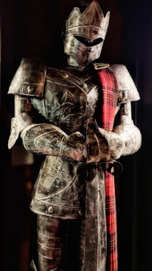 A full suit of medieval armor is displayed, featuring intricate engravings and a red plaid sash draped over the shoulder. The armor includes a helmet with a visor, breastplate, and gauntlets. It stands upright in a slightly darkened setting, giving an imposing and majestic appearance.
