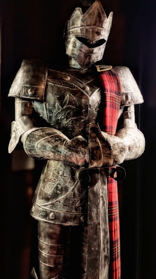A full suit of medieval armor is displayed, featuring intricate engravings and a red plaid sash draped over the shoulder. The armor includes a helmet with a visor, breastplate, and gauntlets. It stands upright in a slightly darkened setting, giving an imposing and majestic appearance.