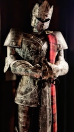 A full suit of medieval armor is displayed, featuring intricate engravings and a red plaid sash draped over the shoulder. The armor includes a helmet with a visor, breastplate, and gauntlets. It stands upright in a slightly darkened setting, giving an imposing and majestic appearance.