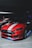 A sports car with a sleek design, painted in vibrant red with a prominent white racing stripe along the hood and roof. The car is parked in a dimly lit indoor garage, with a polished finish that reflects the ambient lighting. The vehicle's headlights and LED accent lights give off a slight glow, adding to its modern aesthetic. Another dark-colored car is slightly visible next to it.