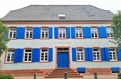 A two-story building with symmetrical architecture features a light gray facade and bright blue shutters on every window. The entrance has a blue door, and the building's name, 'St. Josefsheim', is displayed above the door. Trim and window details are in a reddish-brown color. Trees flank both sides of the building, adding greenery to the scene.