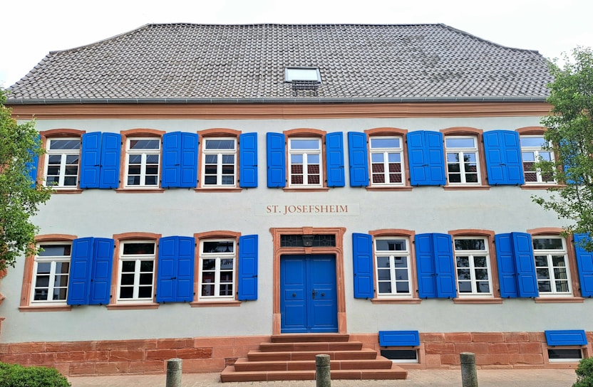 A two-story building with symmetrical architecture features a light gray facade and bright blue shutters on every window. The entrance has a blue door, and the building's name, 'St. Josefsheim', is displayed above the door. Trim and window details are in a reddish-brown color. Trees flank both sides of the building, adding greenery to the scene.